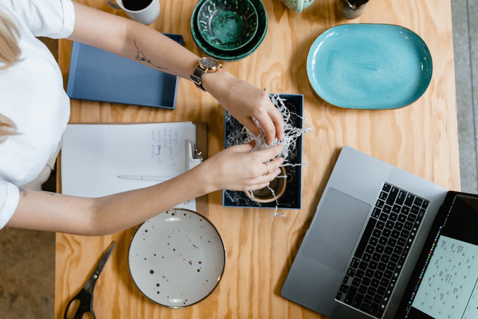 Overhead view of woman's hands preparing a package at a creative workspace with laptop and stationery.