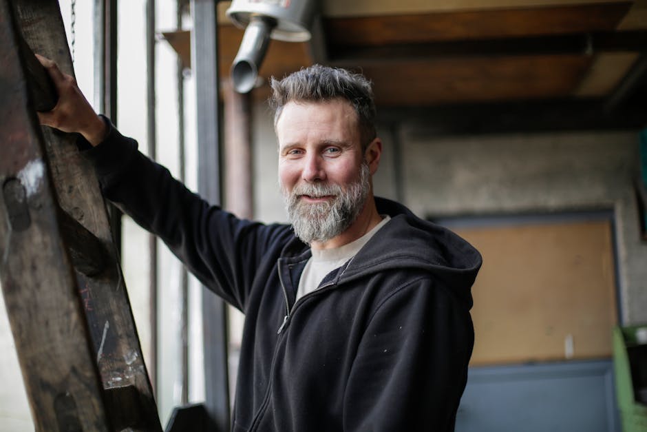 Portrait of a smiling bearded man in a casual hoodie standing indoors with industrial background.