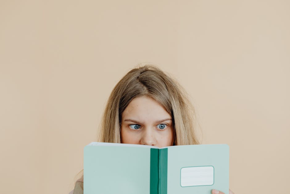Teen girl with blonde hair reading a green notebook, focused and curious.