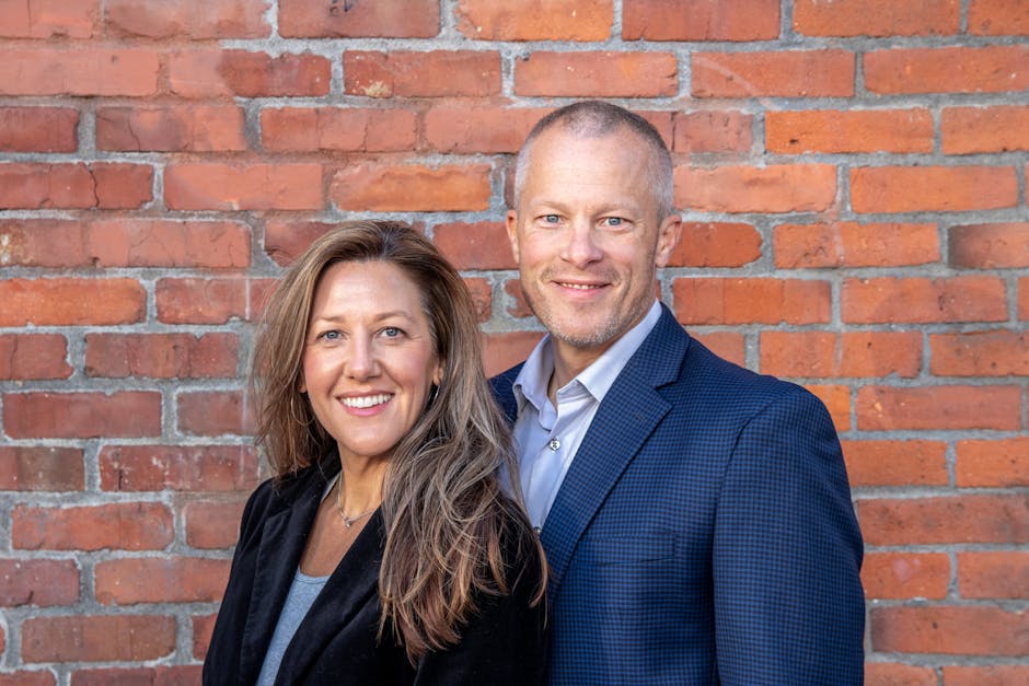 A cheerful middle-aged man and woman in business attire posing against a brick wall, depicting professionalism and partnership.