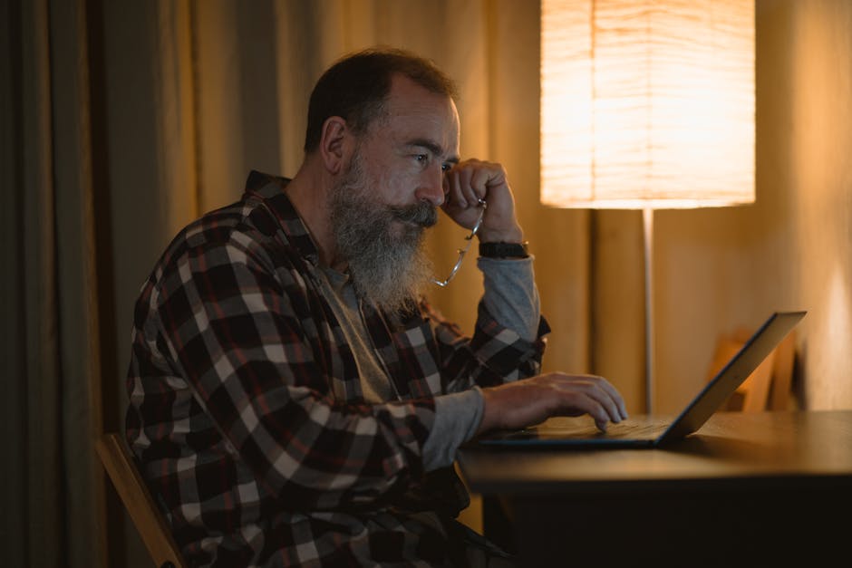 Elderly man with a beard working late on laptop under warm lamp light indoors.