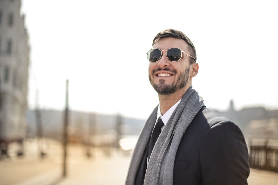 A happy man in a black suit and sunglasses smiles confidently in the bright daylight.