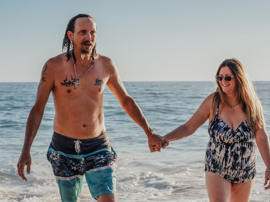 Smiling couple holding hands walking along a sunny beach, enjoying a carefree summer day.