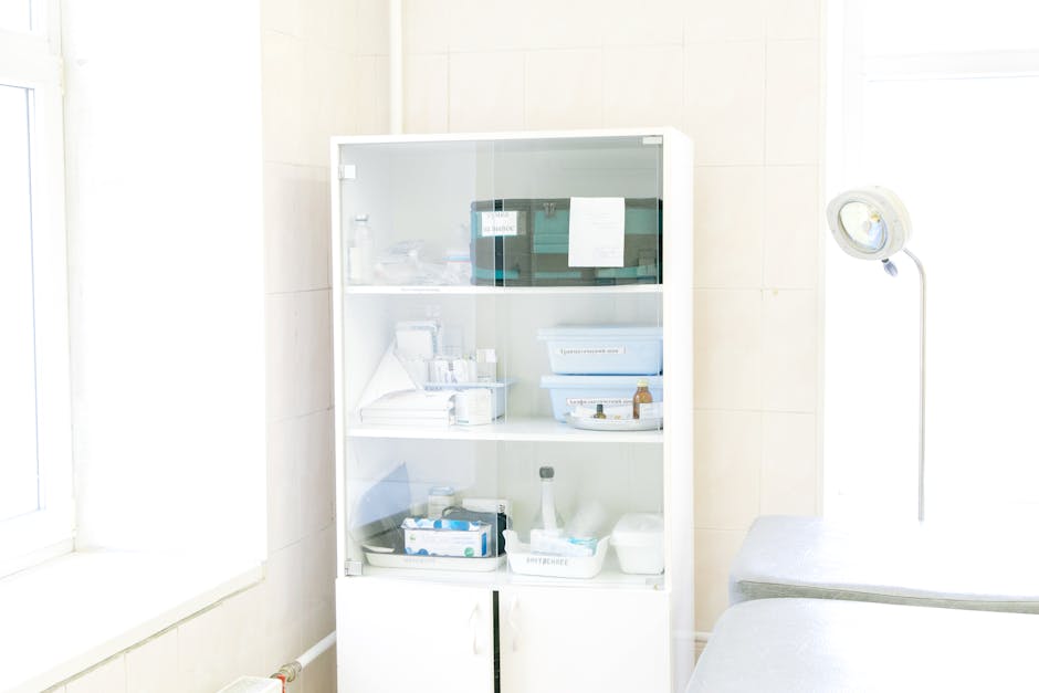 Modern white cabinet with cupboard with various medical instruments and shelves with medical materials near empty white bed near window and lamp in hospital
